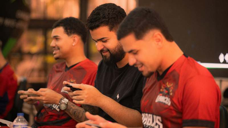 Diverse group of mobile gamers collaborating at a digital table with phones, tablets, and laptops showing multiplayer game interfaces
